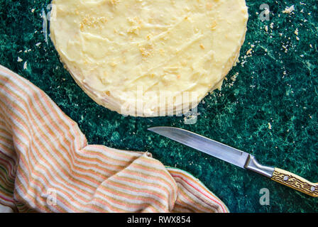 Gâteau rond Napoléon en couches avec de la crème anglaise et saupoudré de miettes, couteau, serviette et mixer sur une table de marbre vert, vue du dessus. Le processus de cuisson, reci Banque D'Images