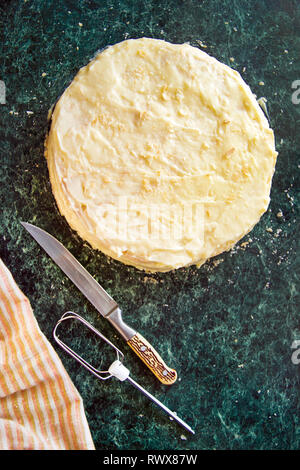 Gâteau rond Napoléon en couches avec de la crème anglaise et saupoudré de miettes, couteau, serviette et mixer sur une table de marbre vert, vue du dessus. Le processus de cuisson, reci Banque D'Images