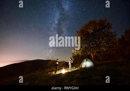 Jeune couple heureux touristes debout à proximité d'un feu allumé tente sous une nuit ciel plein d'étoiles et de Voie lactée. Sur le fond beau ciel étoilé, montagnes et grand arbre Banque D'Images