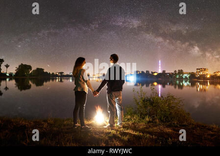 Vue arrière de jeune famille - l'homme et la femme debout sur un lac près de Bonfire, tenant les mains, bénéficiant d'une vue magnifique de nuit ciel plein d'étoiles et Voie lactée au-dessus de l'eau et la ville encore lumineux Banque D'Images