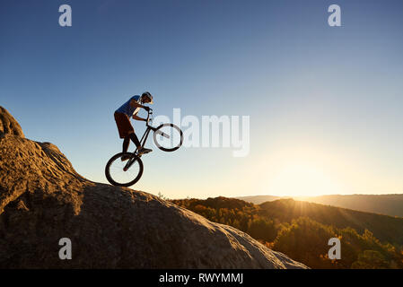 Silhouette de cycliste professionnelle équestre sur roue arrière sur la moto de trial. Sportsman audacieux Biker décisions acrobatic stunt sur le bord de big boulder au coucher du soleil. Concept de sport extrême de vie actif Banque D'Images