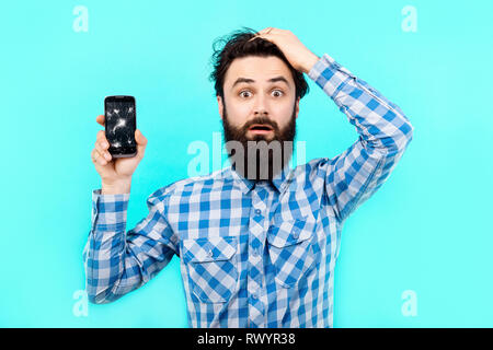 Homme barbu bouleversé à perte avec des téléphones mobiles sur un fond bleu, recherche concept service gsm Banque D'Images