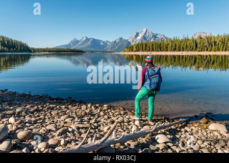 Jeune femme avec sac à dos debout sur la rive, reflétée dans les montagnes, lac, Colter Bay Lake Jackson, chaîne Teton Banque D'Images