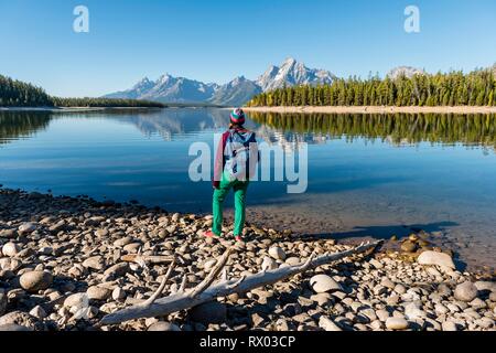 Jeune femme avec sac à dos debout sur la rive, reflétée dans les montagnes, lac, Colter Bay Lake Jackson, chaîne Teton Banque D'Images