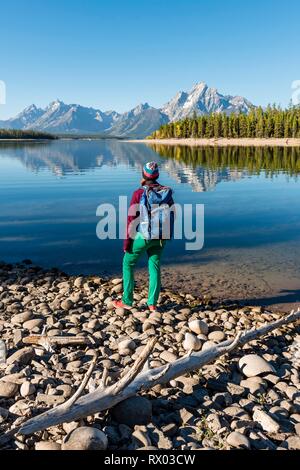 Jeune femme avec sac à dos debout sur la rive, reflétée dans les montagnes, lac, Colter Bay Lake Jackson, chaîne Teton Banque D'Images