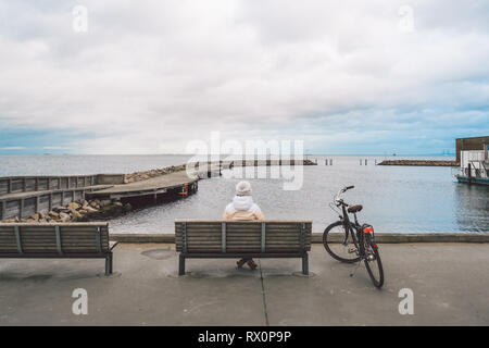 Une jeune femme de race blanche est assis avec son dos sur un banc en bois donnant sur la mer Baltique sur le front de mer à Copenhague au Danemark en hiver en blé nuageux Banque D'Images