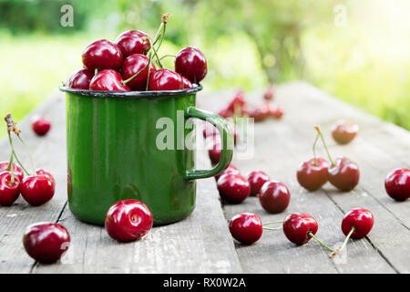 Mug émaillé vert rouge plein de cerises mûres sur banc en bois dans le jardin d'été. Banque D'Images