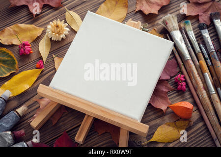 Toile sur chevalet, pinceaux, tubes de peinture pour la peinture et les feuilles d'automne sur le vieux bureau en bois. Vue d'en haut. Mise à plat. Tons rétro. Banque D'Images