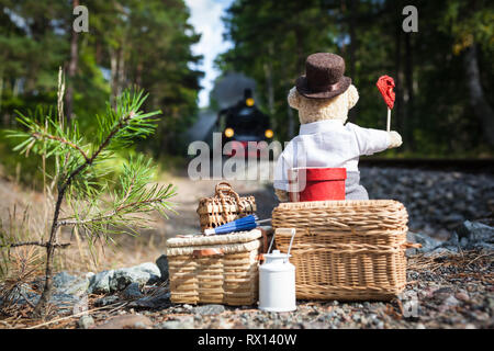 Nounours nostalgique avec gros bagages en attente à l'approche de la voie de train à vapeur, en agitant le drapeau de signal rouge Banque D'Images