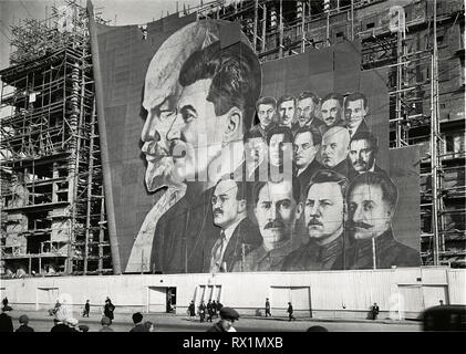 Une grande affiche avec des portraits des dirigeants politiques russes Vladimir Lénine, Joseph Staline et d'autres pour la parade du jour de mai pend sur l'échafaudage d'un bâtiment en construction, Moscou, Russie, 1934.(Photo de Burton Holmes) Banque D'Images