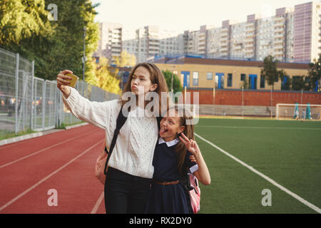 Deux sœurs adolescentes mignonnes prendre vos autoportraits et se tenir sur une aire de jeux. Fille plus âgée est holding cell phone et est plus jeune en serrant les épaules. Piste de course et de f Banque D'Images