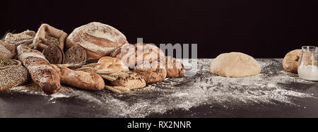 Préparation du pain rustique concept avec pile de pains fraîchement préparés et de pain et de pâte crue sur la surface de la table farinée. Vue panoramique bannière contre bac noir Banque D'Images