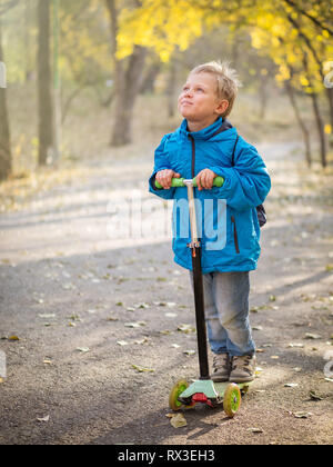 Un garçon en bleu vêtements équitation avec un sourire d'un scooter dans le parc de l'automne. Les rayons de soleil éclairer son visage. Banque D'Images
