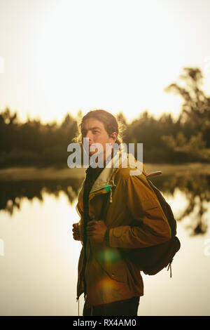 Portrait d'un voyageur debout à côté d'un lac à la route. Veste homme portant un sac à dos et à l'extérieur permanent avec le soleil en arrière-plan. Banque D'Images