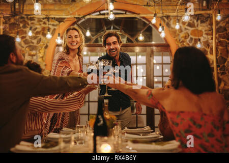 Accueillir couple toasting drinks avec l'invité à dîner. Les jeunes gens joyeux de célébrer avec des boissons au parti. Banque D'Images