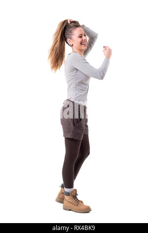 Vue latérale sur la franchise jeune élégant hipster girl holding cheveux en queue et rire avec les yeux fermés. Corps plein isolé sur fond blanc. Banque D'Images