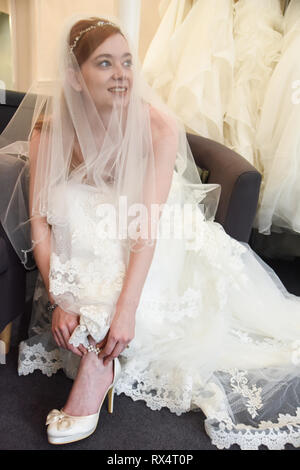 Une jeune fille aux cheveux rouge retty tente sur des robes de mariée dans une boutique. Elle est heureuse et souriante Banque D'Images