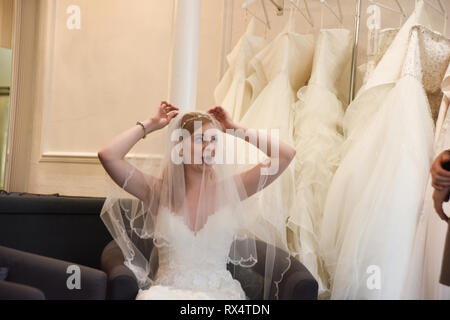 Une jeune fille aux cheveux rouge retty tente sur des robes de mariée dans une boutique. Elle est heureuse et souriante Banque D'Images