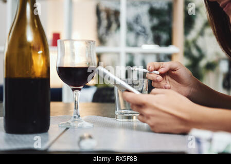 Femme est titulaire d'une cigarette dans une main, un téléphone dans l'autre. À l'aide de verre vide comme un cendrier. Une bouteille et un verre de vin rouge sur la table. Banque D'Images