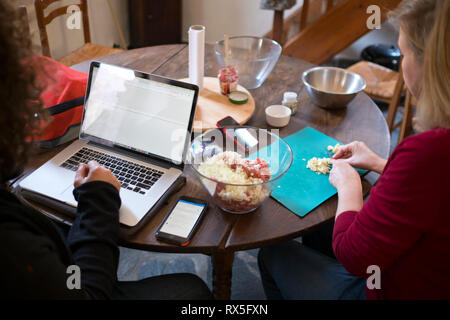 regardant sur les épaules de deux personnes assis à une table en bois, tandis que l'homme travaille sur un ordinateur portable et la femme prépare la nourriture Banque D'Images