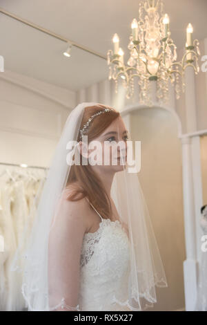 Une jeune fille aux cheveux rouge retty tente sur des robes de mariée dans une boutique. Elle est heureuse et souriante Banque D'Images