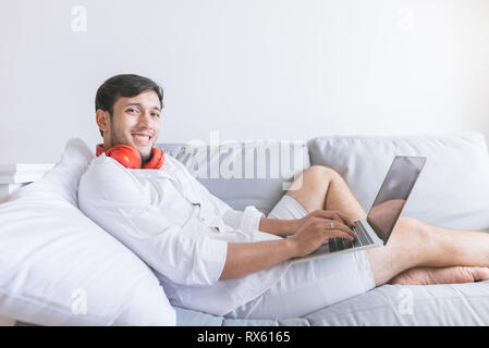 Jeune homme se détendre sur le canapé avec un ordinateur portable et un casque . Vue latérale du bel homme à l'aide de son ordinateur portable et à la recherche à l'appareil photo avec sourire tout en sitti Banque D'Images