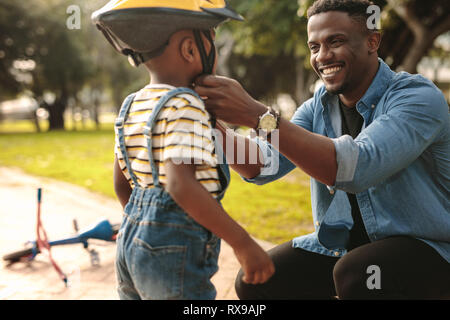 Smiling man helping son port casque pour vélo au parc. Garçon se préparer par le port de casque de vélo pour commencer la randonnée à vélo. Banque D'Images