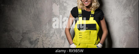 Belle blonde constructeur femme travailleur de construction portant une combinaison jaune travail porter posant contre grunge mur copie espace pour la publicité textuelle Banque D'Images