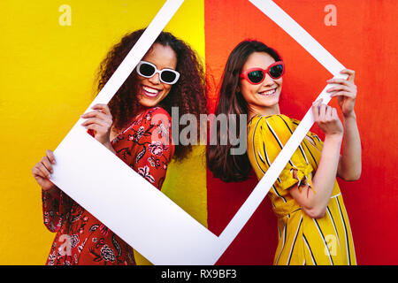 Portrait de belles femmes tenant un cadre photo Blanc dans la main et souriant. Les filles portant des lunettes de se tenir contre un mur de couleur rouge et jaune. Banque D'Images