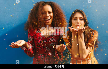 Les jeunes femmes attrayantes soufflant paillettes contre fond bleu. Mixed Race female friends having fun avec des paillettes. Banque D'Images