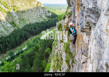 Grimpeur femelle mousquetons de commutation sur une via ferrata itinéraire, mur vertical au-dessus de la vallée, avec une route derrière. Klettersteig vélo à Baia de fie Banque D'Images