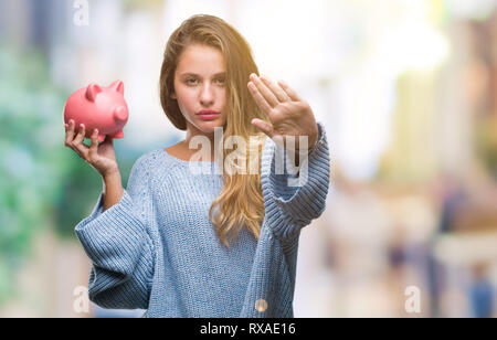 Belle jeune femme blonde holding piggy bank sur fond isolé avec main ouverte faisant stop avec de sérieux et de confiance, la défense de l'expression Banque D'Images