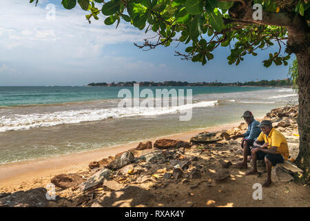 Deux hommes de prendre une pause de la chaleur du soleil de midi et discuter ensemble sous l'ombre d'un arbre sur la plage à Galle, dans le sud de la province de S Banque D'Images