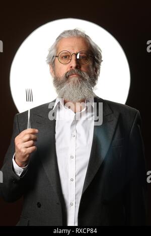 Studio portrait d'un vieil homme aux cheveux gris avec une moustache et barbe hirsute, dans une chemise blanche et veste gris, portant des lunettes et une fourchette, sur un blac Banque D'Images