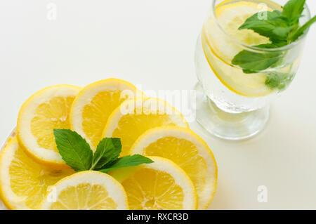 Belle mise en place de verre de limonade froide et la plaque en verre transparent rempli de rondelles de citron avec des feuilles de menthe verte sur fond blanc. Banque D'Images