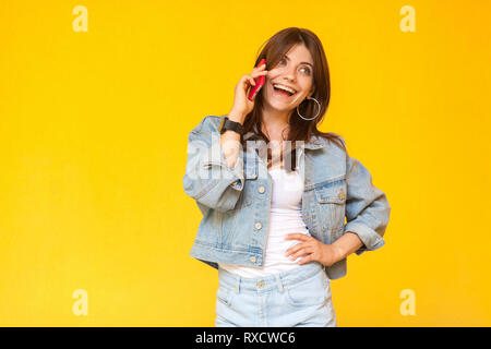 Portrait of happy heureux belle brune jeune femme en denim style casual debout, main sur les côtés, à l'écart et de parler sur son téléphone portable. Banque D'Images