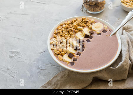 Smoothie bananes avec bol de chocolat, granola et les arachides sur fond de béton gris clair. Le petit-déjeuner végétarien sain. Banque D'Images