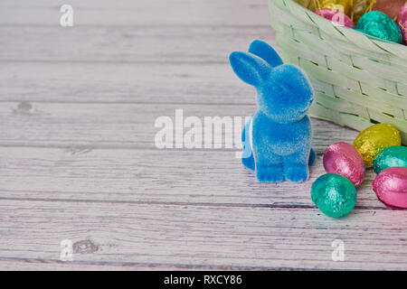 Lapin de Pâques bleu à côté des œufs en chocolat, de couleur en arrière-plan un panier de Pâques sur fond de bois blanc Banque D'Images