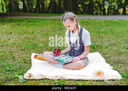 En étudiant l'alimentation uniforme une pomme rouge pour le déjeuner. lycéenne assis sur une couverture dans un parc avec des livres et à faire leurs devoirs Banque D'Images