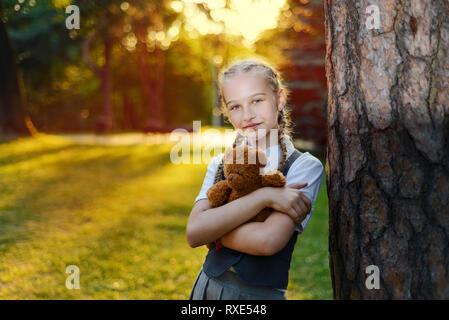 Beau portrait de l'écolière avec des tresses en uniforme au coucher du soleil. l'étudiant se tient près d'un arbre et est titulaire d'un soft toy ours Banque D'Images