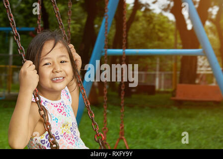 Happy smiling asian petite fille accrocher sur des balançoires en arrière-plan du parc Banque D'Images