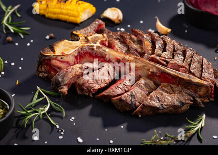 T-bone steak d'aloyau grillé est tranché sur un morceau avec maïs grillé, sauce, romarin, piment, sel, ail sur fond noir. style rustique, haut Banque D'Images