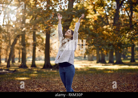 Joyful woman throwing les feuilles d'automne dans l'air au-dessus de sa tête avec un sourire heureux à l'extérieur dans un parc avec des feuilles d'automne colorés sur t Banque D'Images