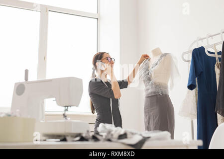 Couturière, tailleur, la mode et concept de montre - belle jeune designer parle sur le téléphone mobile et souriant tout en examinant son travail en Banque D'Images