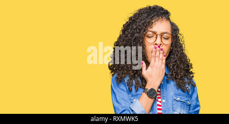 Belle jeune femme aux cheveux bouclés portant des lunettes ennuyer le bâillement fatigué couvrant la bouche avec la main. Agité et somnolence. Banque D'Images