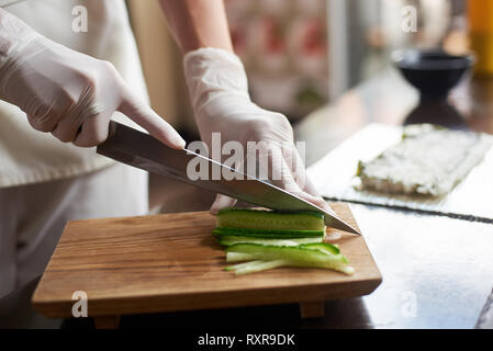 Vue en gros plan de préparation de délicieux sushis roulant dans un restaurant. Les mains dans les gants jetables de trancher le concombre sur planche de bois. Banque D'Images