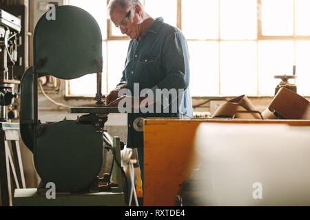 Travailleur senior travaillant sur machine à bande dans son atelier. Menuisier bois de coupe sur la machine à l'atelier de menuiserie. Banque D'Images