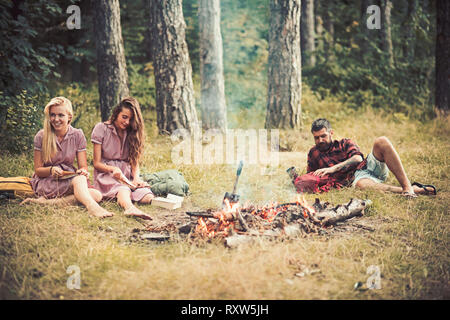 Les filles assis près d'un feu de camp alors que la lecture de livres. Barbu allongé sur l'herbe dans la forêt. Amis camping en été Banque D'Images