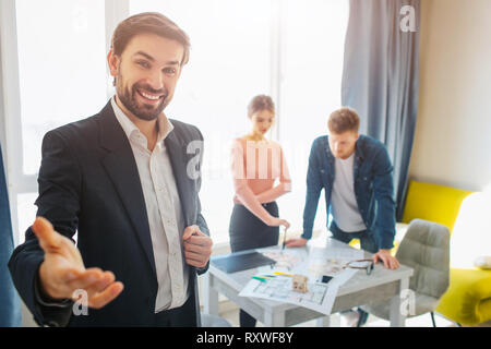 Couple acheter ou louer vacances ensemble. Cheerful realtor barbus atteignent à l'appareil photo à la main et sourire. Jeune couple se tenir derrière à table et regarder Banque D'Images