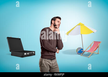 Penser l'homme avec valise ouverte, chaise de plage, ballon et parapluie sur fond bleu. Banque D'Images